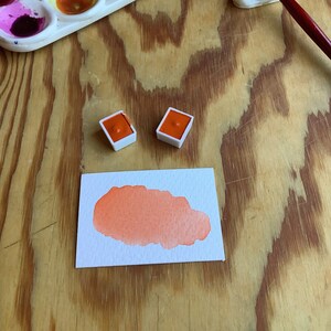 May include: A watercolor painting of a peachy orange color on a white paper card. Two small orange watercolor paint pans are on a wooden surface next to the card. A paintbrush and a paintbrush holder are also on the surface.