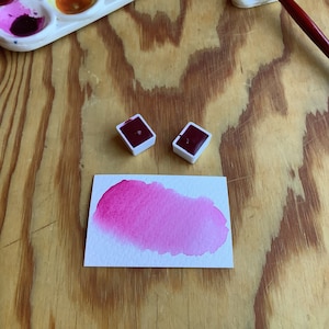 May include: A watercolor paint set with a tray of assorted colors, two small pans of magenta paint, and a brush resting on a white holder. A pink watercolor wash is painted on a white paper.