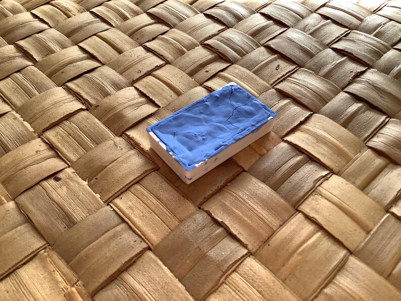 May include: A single blue watercolor paint pan on a woven straw surface.