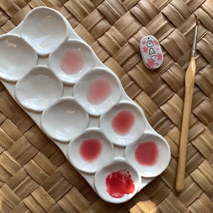 Pu&ograve; includere: Una tavolozza di pittura in ceramica bianca con nove pozzetti, riempiti con tonalit&agrave; di vernice rosa e rossa. Un piccolo cartellino in ceramica con la parola "GAY" in lettere bianche &egrave; visibile sullo sfondo. &Egrave; visibile anche un pennello di legno.