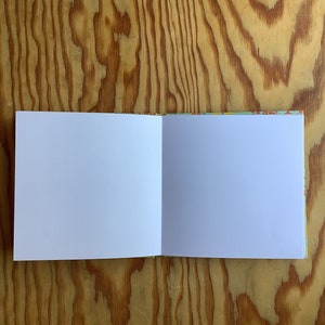 Puede incluir: Un cuaderno abierto en blanco con una cubierta blanca y una cubierta con un patr&oacute;n colorido. El cuaderno est&aacute; abierto sobre una superficie de madera.