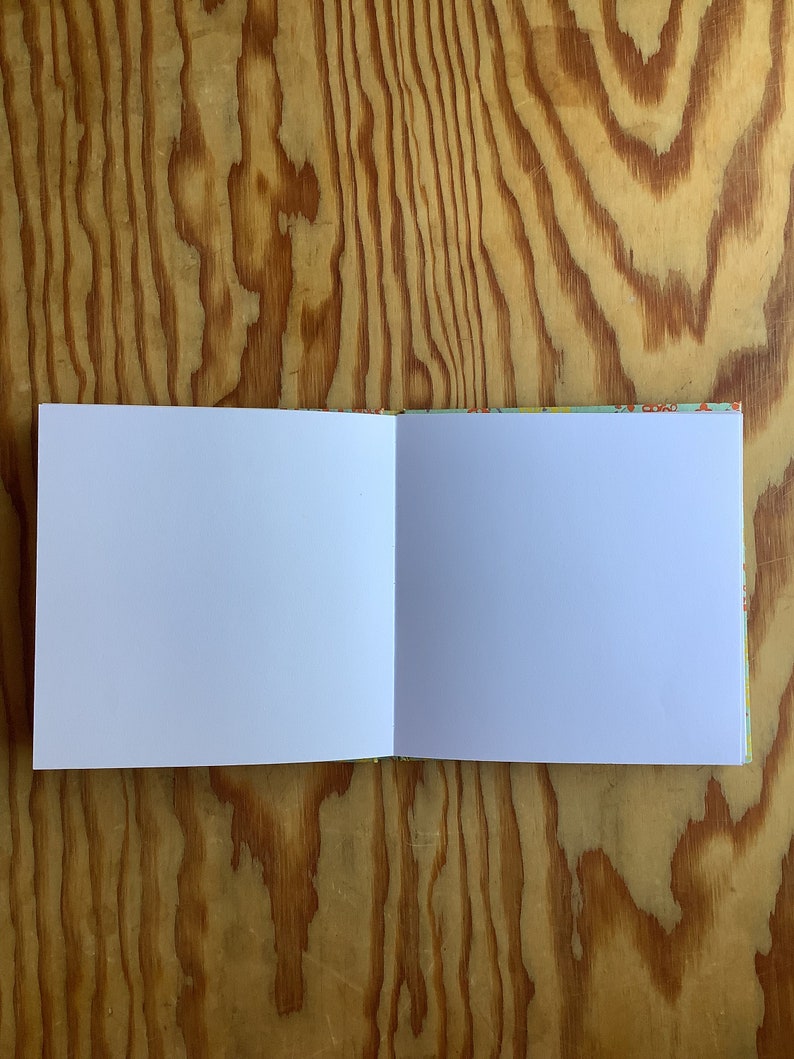 Puede incluir: Un cuaderno abierto en blanco con una cubierta blanca y una cubierta con un patr&oacute;n colorido. El cuaderno est&aacute; abierto sobre una superficie de madera.