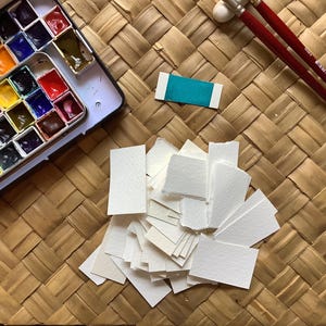 Può includere: Un set di acquerelli in una custodia di plastica con una varietà di colori. Ci sono anche diversi piccoli pezzi di carta bianca e un piccolo pezzo di carta turchese.