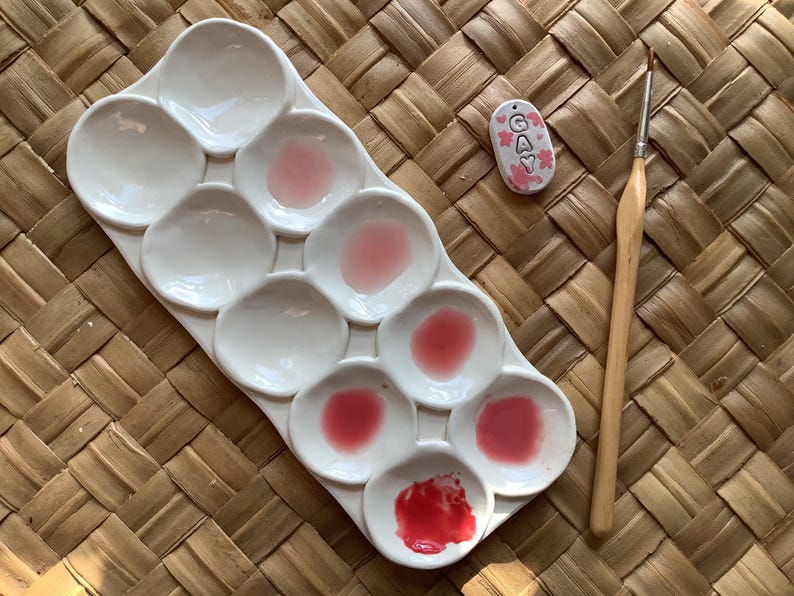 Pu&ograve; includere: Una tavolozza di pittura in ceramica bianca con nove pozzetti, riempiti con tonalit&agrave; di vernice rosa e rossa. Un piccolo cartellino in ceramica con la parola "GAY" in lettere bianche &egrave; visibile sullo sfondo. &Egrave; visibile anche un pennello di legno.