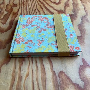 Puede incluir: Un peque&ntilde;o cuaderno azul con un patr&oacute;n floral en tonos naranja, amarillo y morado. El cuaderno tiene un cierre de banda el&aacute;stica marr&oacute;n.