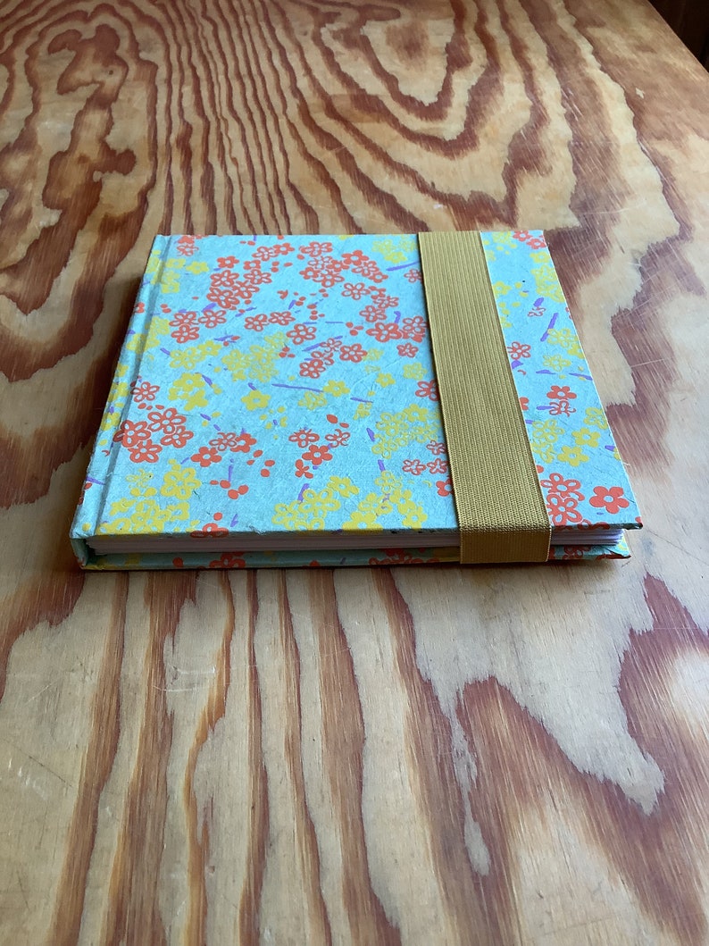Puede incluir: Un peque&ntilde;o cuaderno azul con un patr&oacute;n floral en tonos naranja, amarillo y morado. El cuaderno tiene un cierre de banda el&aacute;stica marr&oacute;n.