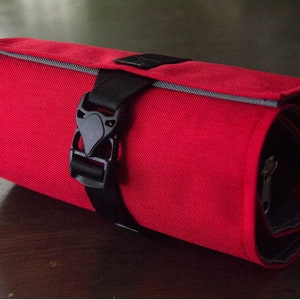 Puede incluir: Una bolsa enrollable de lona roja con una correa negra y un cierre de hebilla. La bolsa está enrollada y asegurada con la correa.