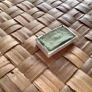 May include: A single watercolor paint pan with a green pigment. The pan is in a white plastic container and is sitting on a woven surface.