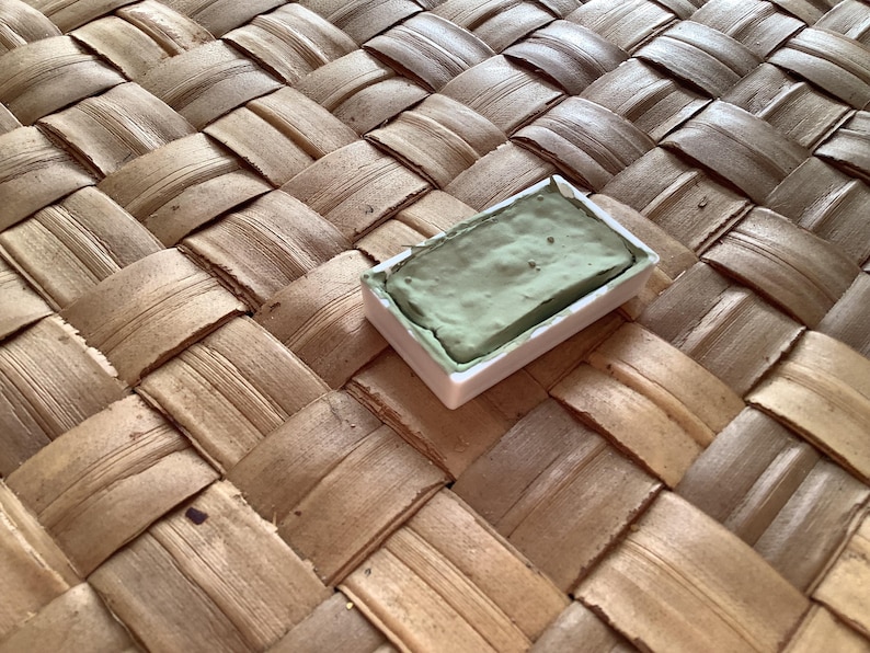 May include: A single watercolor paint pan with a green pigment. The pan is in a white plastic container and is sitting on a woven surface.
