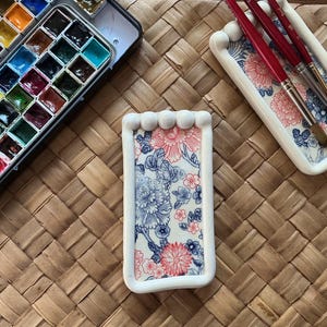 May include: A rectangular white ceramic brush rest featuring a floral pattern in red and blue, positioned next to a metal watercolour paint set and a matching ceramic tray holding two paintbrushes with red handles and brown bristles.