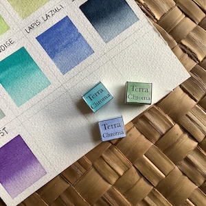 Op de afbeelding: Drie aquarelverfpannetjes met het label "Terra Chroma" in blauw, groen en lichtblauw. De pannetjes liggen op wit aquarelpapier met kleurstalen.