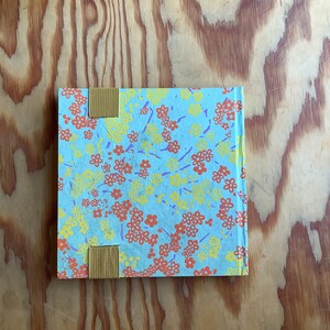 Puede incluir: Un peque&ntilde;o cuaderno azul con un patr&oacute;n floral en tonos amarillos, naranjas y morados. El cuaderno tiene dos marcadores de cinta amarilla.