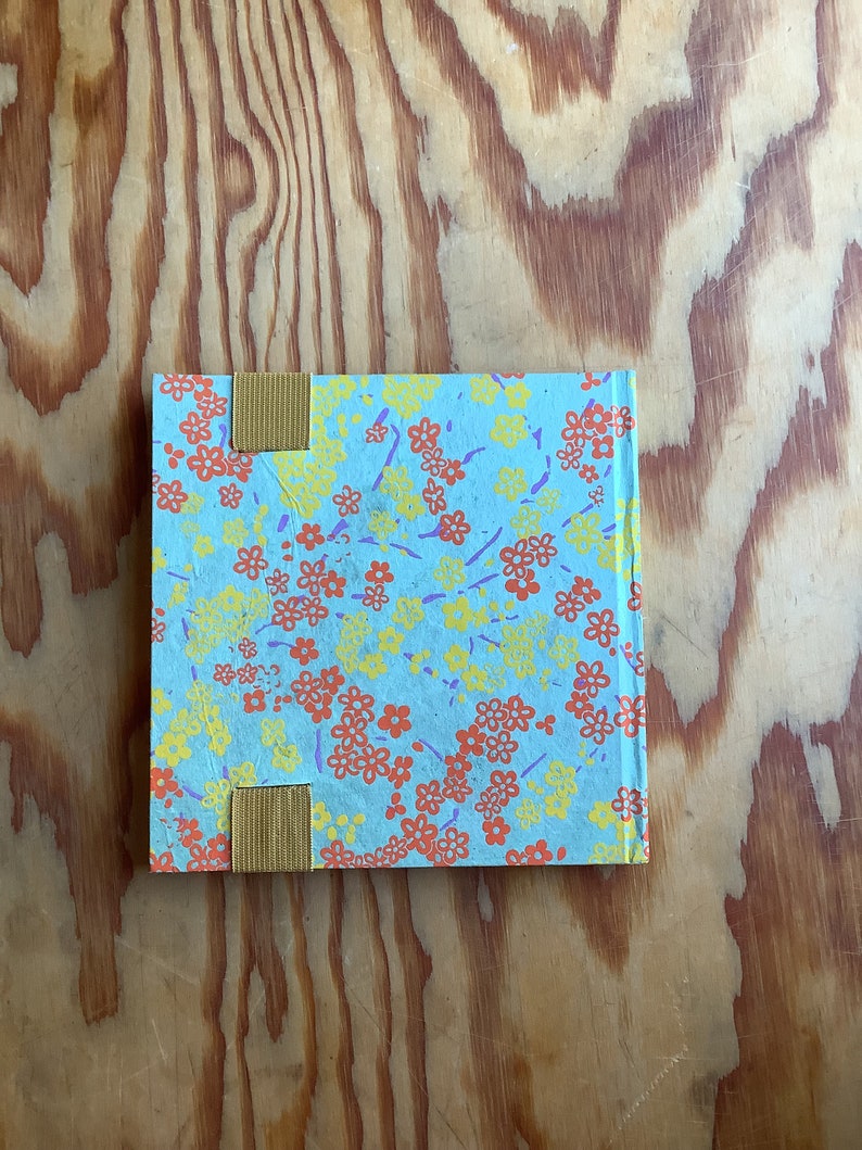 Puede incluir: Un peque&ntilde;o cuaderno azul con un patr&oacute;n floral en tonos amarillos, naranjas y morados. El cuaderno tiene dos marcadores de cinta amarilla.