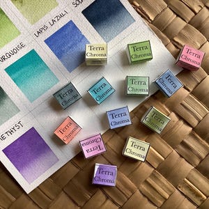 Puede incluir: Una colección de 12 pastillas de pintura de acuarela, cada una etiquetada como "Terra Chroma". Las pastillas están dispuestas en una cuadrícula sobre una superficie tejida. Las pastillas de pintura son de varios colores, incluyendo verde, azul, rosa y morado.