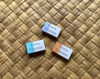 Può includere: Tre scatole rettangolari con il testo "Cerra Chroma". Ogni scatola presenta una diversa sfumatura di colore: blu e bianco, blu e arancione e arancione e bianco. Sono su una superficie intrecciata marrone chiaro.