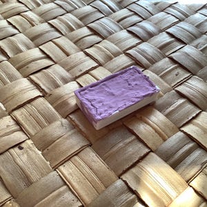 Pu&ograve; includere: Un piccolo blocco rettangolare di acquerello viola. La vernice &egrave; in una vaschetta bianca con un bordo leggermente rialzato. La vaschetta &egrave; appoggiata su una superficie intrecciata di colore marrone chiaro.