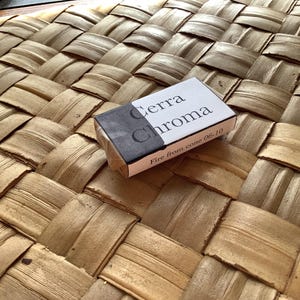 May include: A small white box with black lettering that reads "Terra Chroma" and "Fire from cone 06-10". The box is sitting on a woven brown surface.
