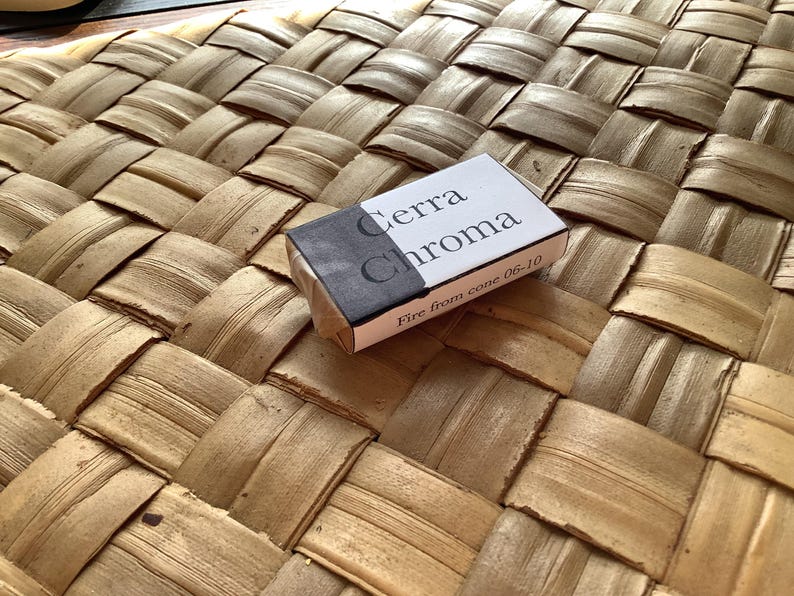 May include: A small white box with black lettering that reads "Terra Chroma" and "Fire from cone 06-10". The box is sitting on a woven brown surface.