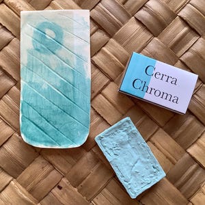 Pu&ograve; includere: Un campione di vernice ad acquerello blu turchese su una piastrella di ceramica bianca, un piccolo blocco di vernice blu turchese e una scatola bianca con il testo "Cerra Chroma" stampato su di essa.
