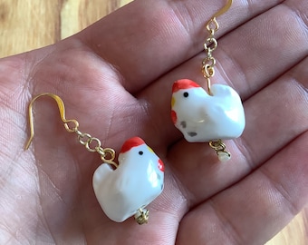 Pode incluir: Um par de brincos de galinha em cerâmica branca com detalhes em vermelho e amarelo. Os brincos têm ganchos e elos de corrente dourados. As galinhas são pequenas e têm um design caprichoso.