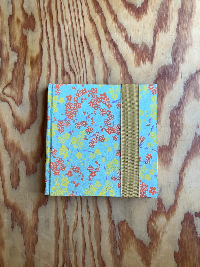 Puede incluir: Un peque&ntilde;o cuaderno azul con un patr&oacute;n floral en tonos rojos, amarillos y morados. El cuaderno tiene un cierre el&aacute;stico marr&oacute;n.