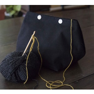 Può includere: Una borsa in tela nera con due bottoni a pressione argentati e un gomitolo di lana giallo con un uncinetto di legno.