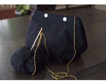 May include: A black canvas bag with two silver snaps and a yellow yarn ball with a wooden crochet hook.