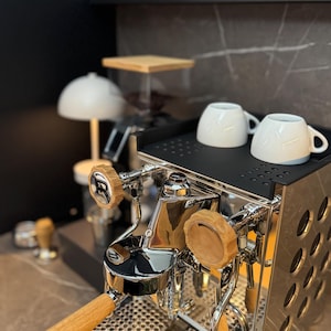 Könnte beinhalten: Nahaufnahme einer silbernen Espressomaschine mit Holzgriffen und zwei weißen Keramikbechern auf einem schwarzen Tablett.