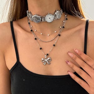 Könnte beinhalten: Eine mehrlagige silberne Halskette mit einem Schleifenanhänger und einer Choker-Halskette aus mehreren verbundenen Zifferblättern.