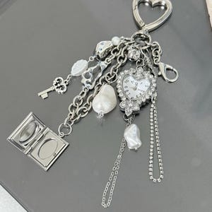Peut inclure: Un porte-clés en argent avec un charme en forme de cœur avec une montre, un médaillon, des clés, des perles et une chaîne. Le cadran de la montre affiche des chiffres romains et le mot "BOER" imprimé dessus.