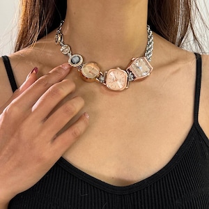 Puede incluir: Un collar de cadena plateada con múltiples caras de reloj pequeñas, redondas y cuadradas unidas. Las caras de los relojes son de color dorado y tienen manecillas negras y marcas de números romanos.