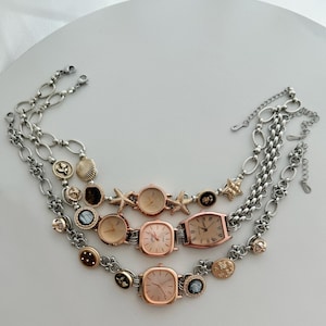 Puede incluir: Tres relojes de tono dorado con caras cuadradas y rectangulares en pulseras de cadena plateada con varios encantos, incluyendo conchas marinas, estrellas y otros elementos decorativos.