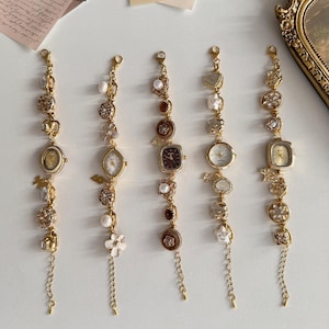 Puede incluir: Cinco pulseras de abalorios de estilo vintage en tono dorado con esferas de reloj. Cada pulsera presenta una forma de esfera diferente y está adornada con perlas, cristales y abalorios decorativos. Las pulseras se exhiben sobre una superficie blanca.