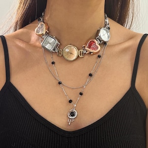 May include: A unique necklace featuring a series of watch faces in various shapes, including square and heart-shaped, connected by a silver chain. The necklace has a layered design with black bead accents and a pendant with a black stone.