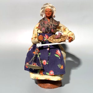 May include: A handmade doll wearing a white lace head covering, a floral patterned dress, and a brown shawl. The doll is holding a basket of flowers and a scale.