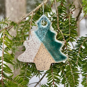 May include: A ceramic Christmas tree ornament with a speckled beige, white, and teal glaze. The tree-shaped ornament is hanging from a silver wire on a branch with green needles. The ornament has a glossy finish.