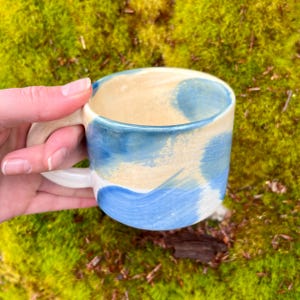 May include: A handmade ceramic mug with a white handle and a blue and yellow glaze. The mug has a unique, abstract design.