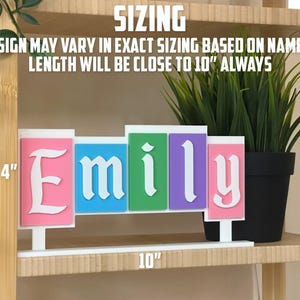 May include: A colourful custom name sign spelling "Emily" in white, with each letter on a different coloured background: pink, blue, green, and purple. The sign is approximately 25 cm long and 10 cm tall. The text "SIZING" is at the top.