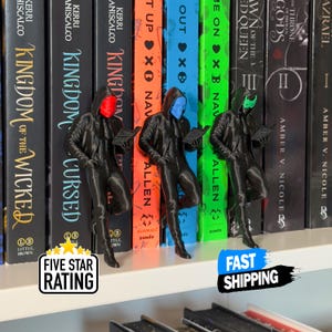 May include: Three black bookends shaped like figures in hooded jackets, each with a different colored face: red, blue, and green. They are leaning against a row of books. The image also includes a "Five Star Rating" and "Fast Shipping" graphic.