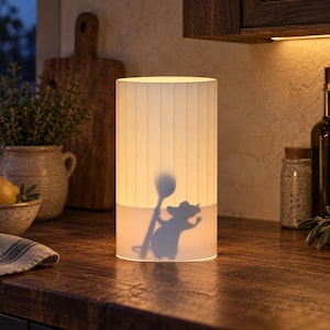 May include: A cylindrical table lamp with a white, vertically striped shade. The lamp emits a warm glow, projecting a dark silhouette of a mouse holding a large wooden spoon onto the lower portion of the shade, creating a distinct visual contrast.
