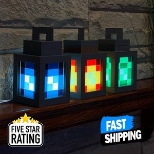 以下が含まれることがあります： 青、赤/黄、緑色のパネルが光る、黒いピクセルスタイルのランタン3つ。各ランタンは正方形のトップとハンドルが付いています。ランタンは、石壁を背景に、暗い木製の表面に展示されています。