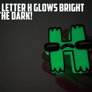 May include: A green glow-in-the-dark letter H with a cartoon face and sunglasses. The letter H glows brightly in the dark. The text "THE LETTER H GLOWS BRIGHT IN THE DARK!" is above the letter.