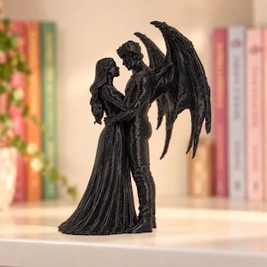 May include: A black figurine depicting two figures standing in an embrace. One figure has long hair and wears a floor-length gown, while the other figure features large, detailed wings extending from the back. The sculpture has a textured surface.