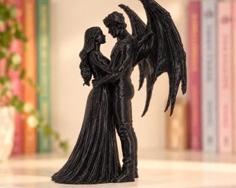 May include: A black figurine depicting two figures standing in an embrace. One figure has long hair and wears a floor-length gown, while the other figure features large, detailed wings extending from the back. The sculpture has a textured surface.