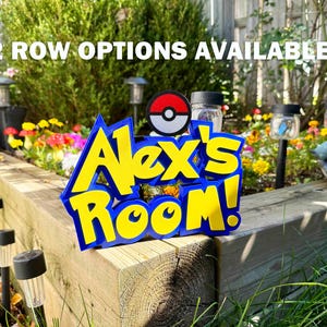 May include: A blue and yellow sign reading "Alex's Room!" with a Pokeball design, set in a garden with flowers and solar lights. The text "2 ROW OPTIONS AVAILABLE!" is above the sign. The sign is made of wood.