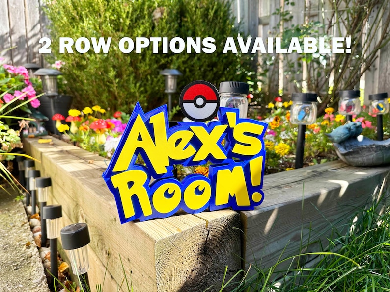 May include: A blue and yellow sign reading "Alex's Room!" with a Pokeball design, set in a garden with flowers and solar lights. The text "2 ROW OPTIONS AVAILABLE!" is above the sign. The sign is made of wood.