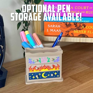 May include: A decorative wooden pen holder with three drawers, featuring floral, flame, and celestial designs. The text "OPTIONAL PEN STORAGE AVAILABLE!" is displayed above. The holder contains colourful pens and a blue pen.