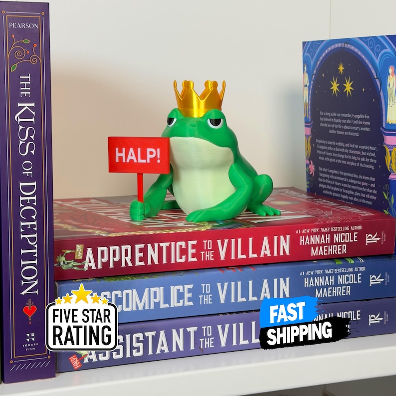 May include: A green frog figurine wearing a gold crown and holding a red sign that says "HALP!" sits atop a stack of books. The books have titles like "Apprentice to the Villain" and "Comprice to the Villain". A "Five Star Rating" sticker is visible.