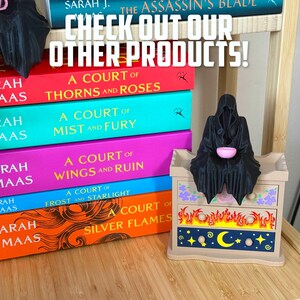 May include: A display of fantasy novels by Sarah J. Maas, including "A Court of Thorns and Roses", with a decorative chest featuring flame and celestial designs. A black figurine sits atop the chest. The text "CHECK OUT OUR OTHER PRODUCTS!" is at the top.