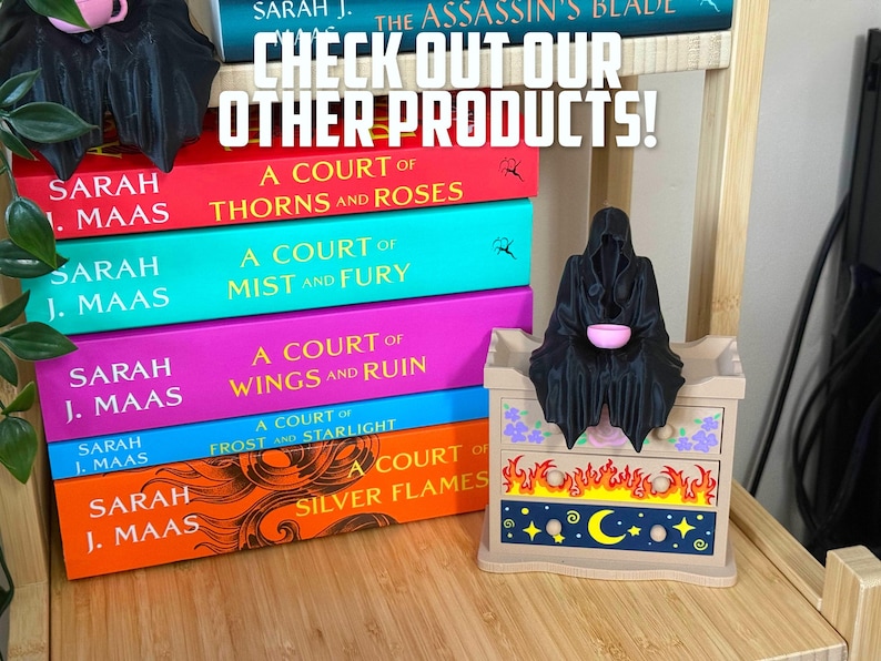 May include: A display of fantasy novels by Sarah J. Maas, including "A Court of Thorns and Roses", with a decorative chest featuring flame and celestial designs. A black figurine sits atop the chest. The text "CHECK OUT OUR OTHER PRODUCTS!" is at the top.